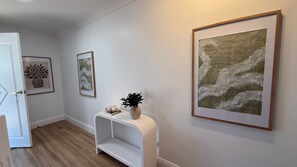 A hallway with a white door, a white console table with a vase and a cushion, and three framed artworks on the wall. - Sage - Rainbow Beach (Rainbow Beach)