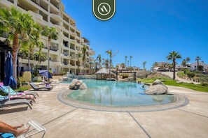 A resort pool area with lounge chairs, palm trees, and a large building in the background.