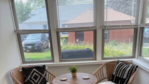 Een veranda met rieten stoelen, een klein tafeltje en uitzicht op een geparkeerde auto en groen buiten.