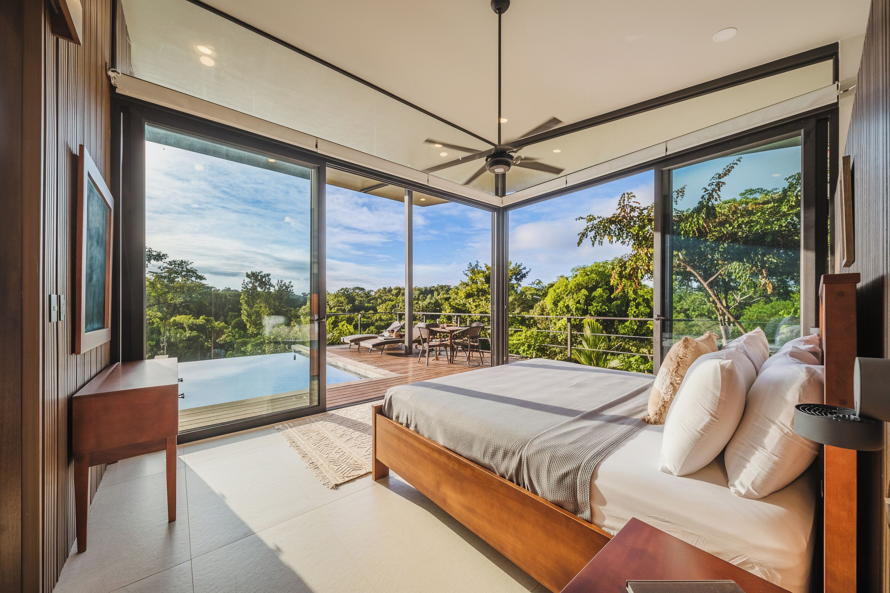 A modern bedroom with a large bed, a ceiling fan, and a view of a pool and lush greenery.