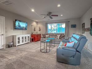 Een woonkamer met een grijze bank, een kleine tafel, een aan de muur bevestigde televisie en een raam met gordijnen.
