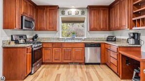A kitchen with wooden cabinets, a granite countertop, a stainless steel refrigerator, a microwave, a stove, a dishwasher, and a window with a view of trees.