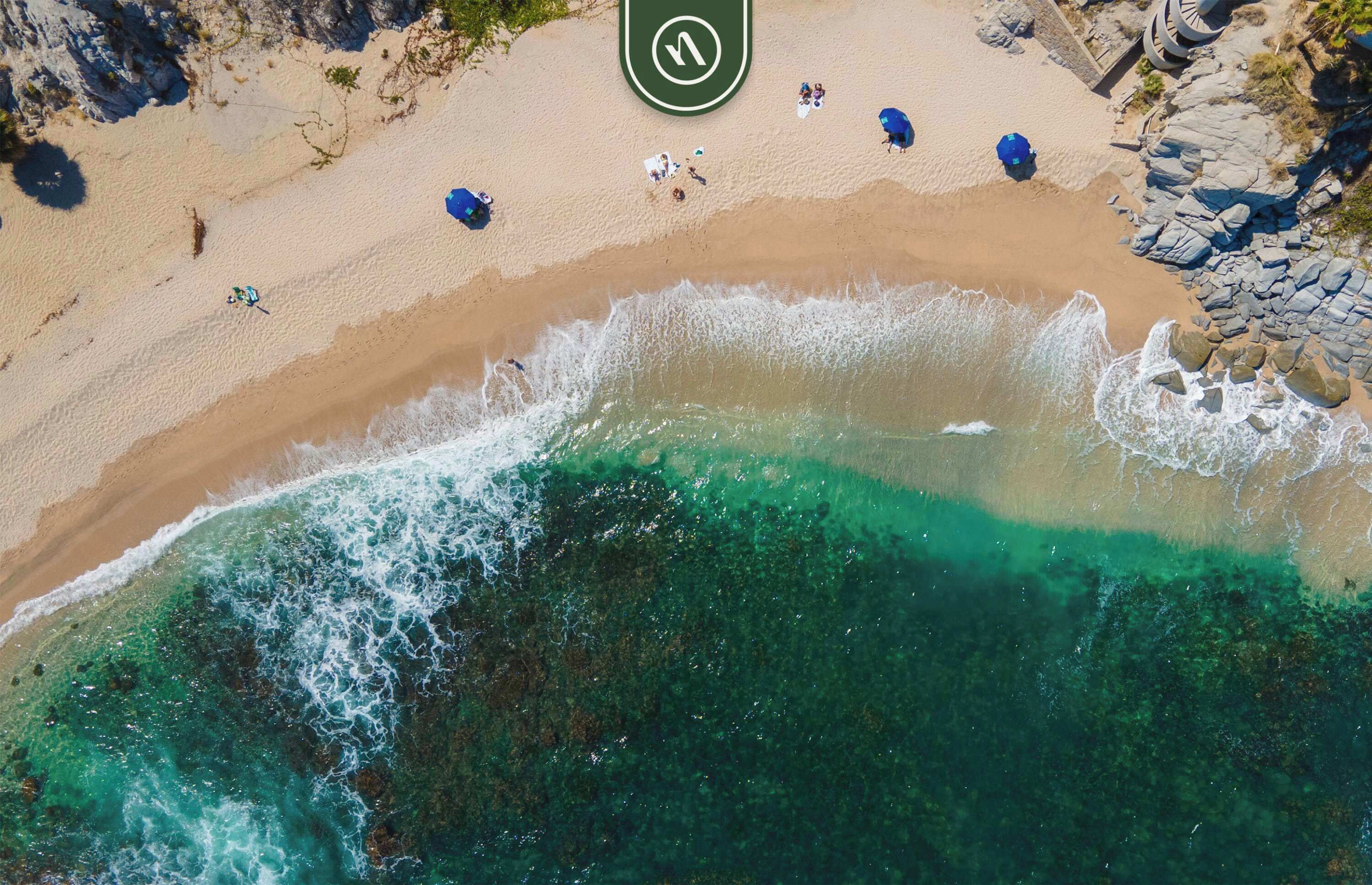 En vy över en strand från ovan, med kristallklart turkost vatten, en sandstrand och några få personer samt parasoller.