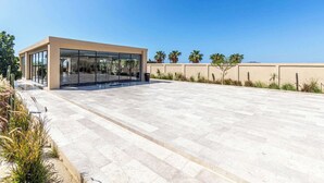 A modern building with a glass facade, a paved patio area, and palm trees in the background. - Luxury 2 BR Condo - Oceanview Pool - Gym (Cabo San Lucas)