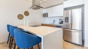 A modern kitchen with a central island, stainless steel appliances, and a seating area with blue chairs. - Luxury 2 BR Condo - Oceanview Pool - Gym (Cabo San Lucas)