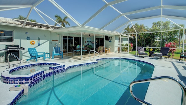 A covered outdoor swimming pool with blue-tiled edges, a patio area with chairs, and a view of the surrounding greenery. - Hot Tub + Heated Pool, Gulf Access, Waterfront with Lanai TV (Cape Coral)