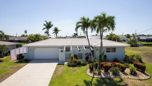 A single-story house with a white exterior, a covered garage, and a well-maintained lawn with palm trees and flowering plants. - Hot Tub + Heated Pool, Gulf Access, Waterfront with Lanai TV (Cape Coral)