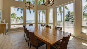 Un comedor espacioso con una gran mesa de madera y sillas, ventanas amplias con vistas a palmeras y un balcón, además de una lámpara colgante.