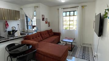 Un salon moderne comprenant un canapé d’angle, une table à manger avec des chaises, un coin cuisine avec une bouilloire et un ventilateur de plafond.