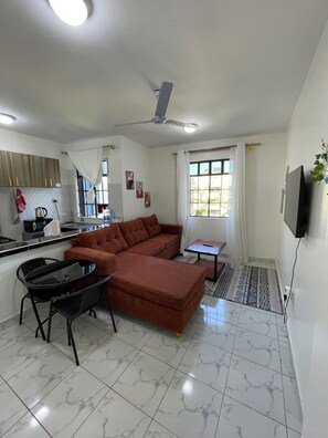 Una sala moderna con un sofá modular, una mesa de comedor con sillas, una zona de cocina con hervidor y un ventilador de techo.