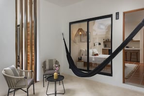 Una habitación acogedora con una hamaca, una silla de mimbre, una mesita redonda y una cama con un ventanal grande.