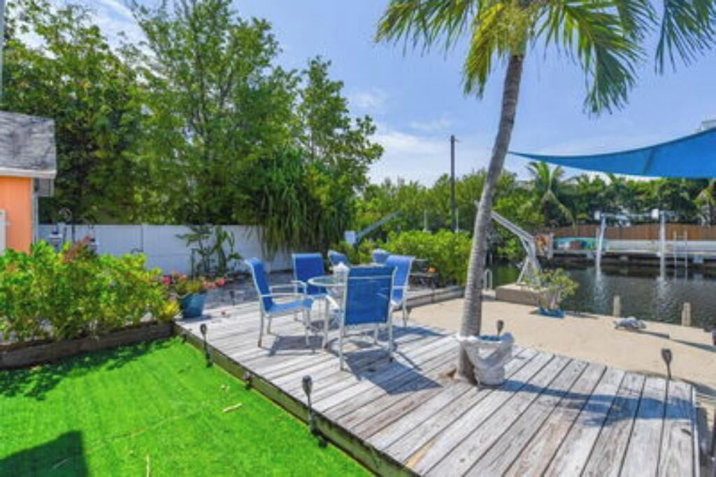 A wooden deck with blue chairs and a table, surrounded by greenery and a body of water.