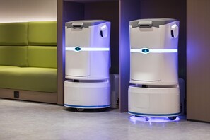 Two robotic vacuum cleaners in a hallway with a green upholstered bench and a wooden door. - Ji Hotel Guangzhou Caihongqiao Metro Station (Guangzhou)
