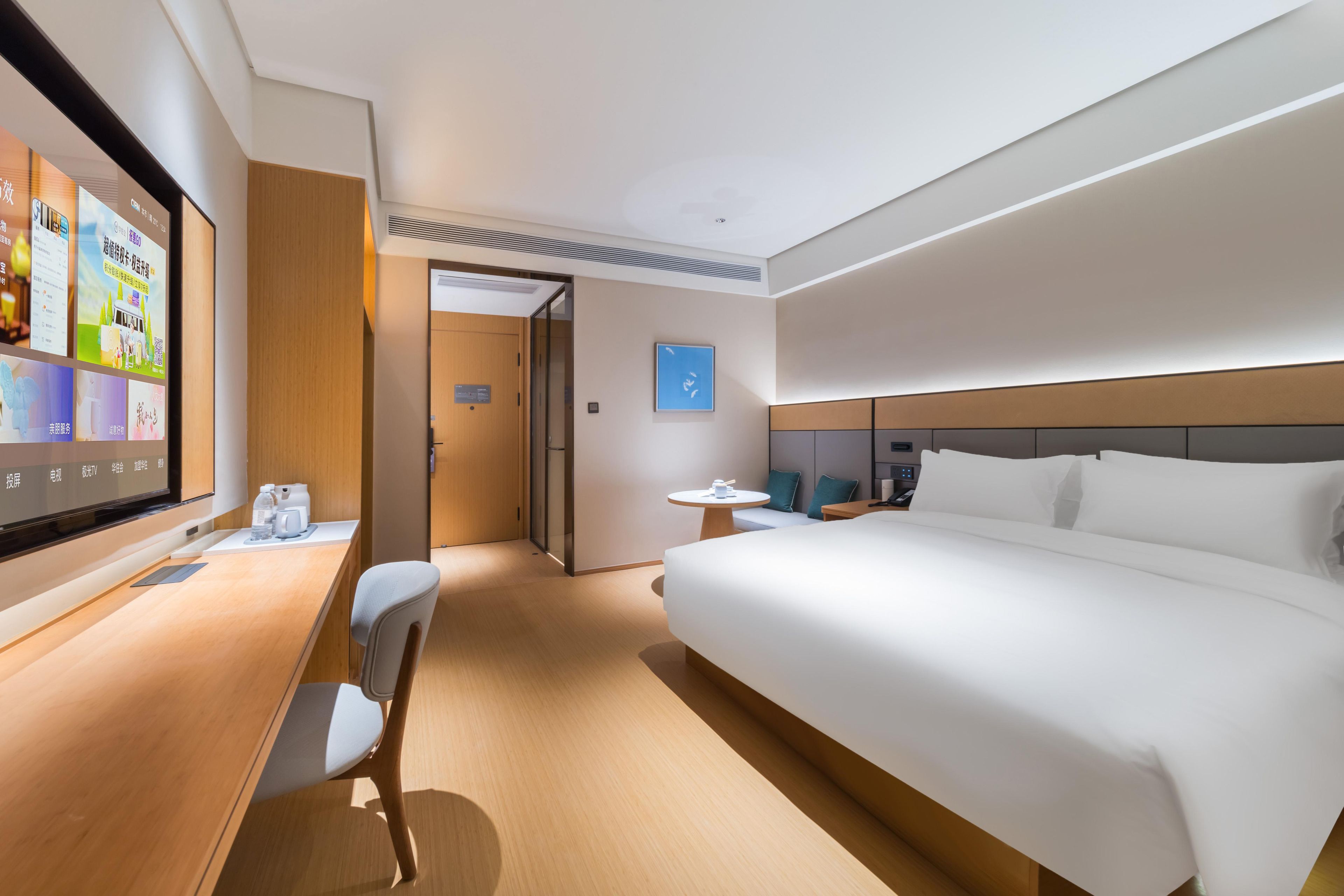 A hotel room with a bed, a desk with a chair, a small table, and a TV displaying a menu.