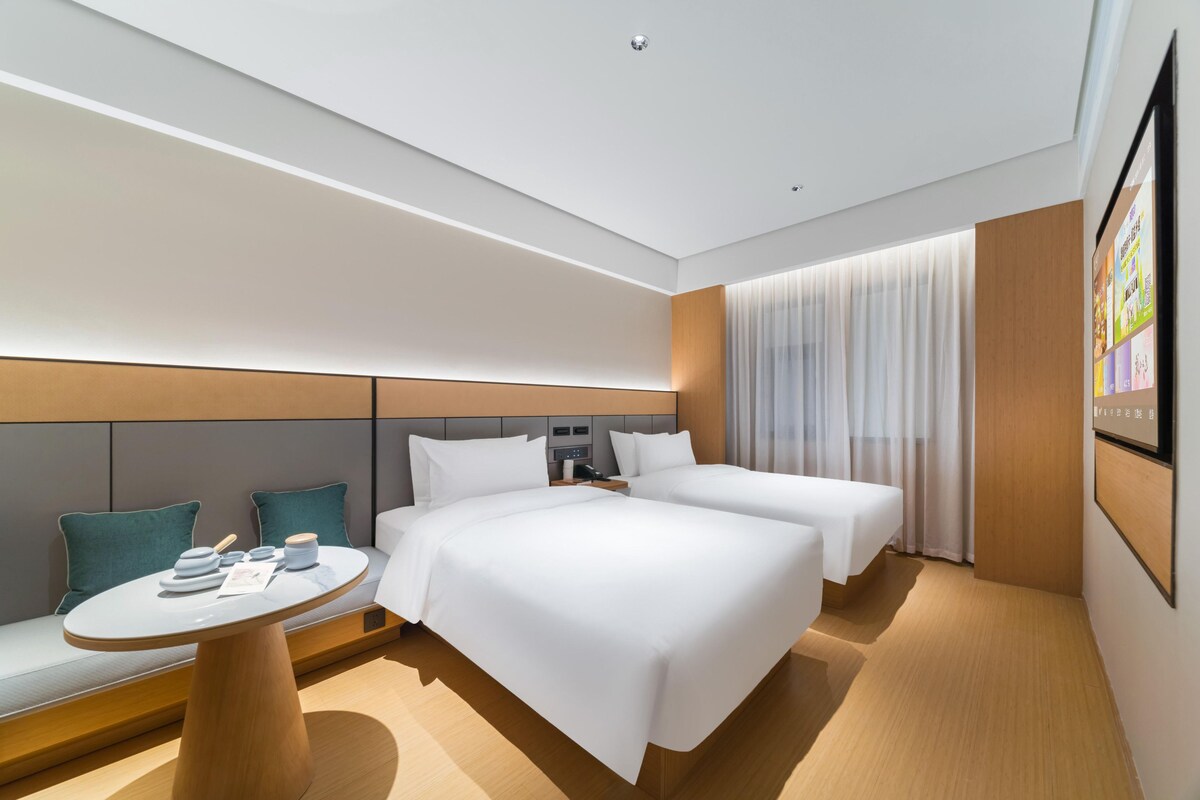 Quarto de hotel moderno com duas camas, uma mesa redonda e um espaço de estar com almofadas.