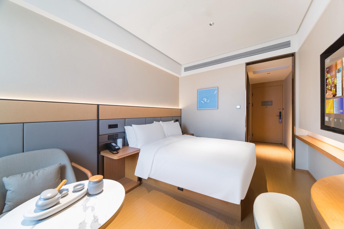 Ein Hotelzimmer mit Bett, Schreibtisch mit Stuhl, einem kleinen Tisch mit Tablett und Fernseher an der Wand.
