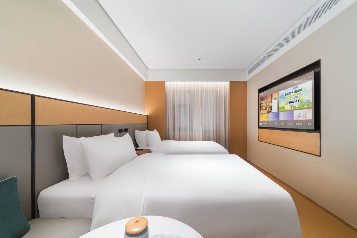 Une chambre d’hôtel avec deux lits, un téléviseur fixé au mur et une fenêtre avec des rideaux légers.