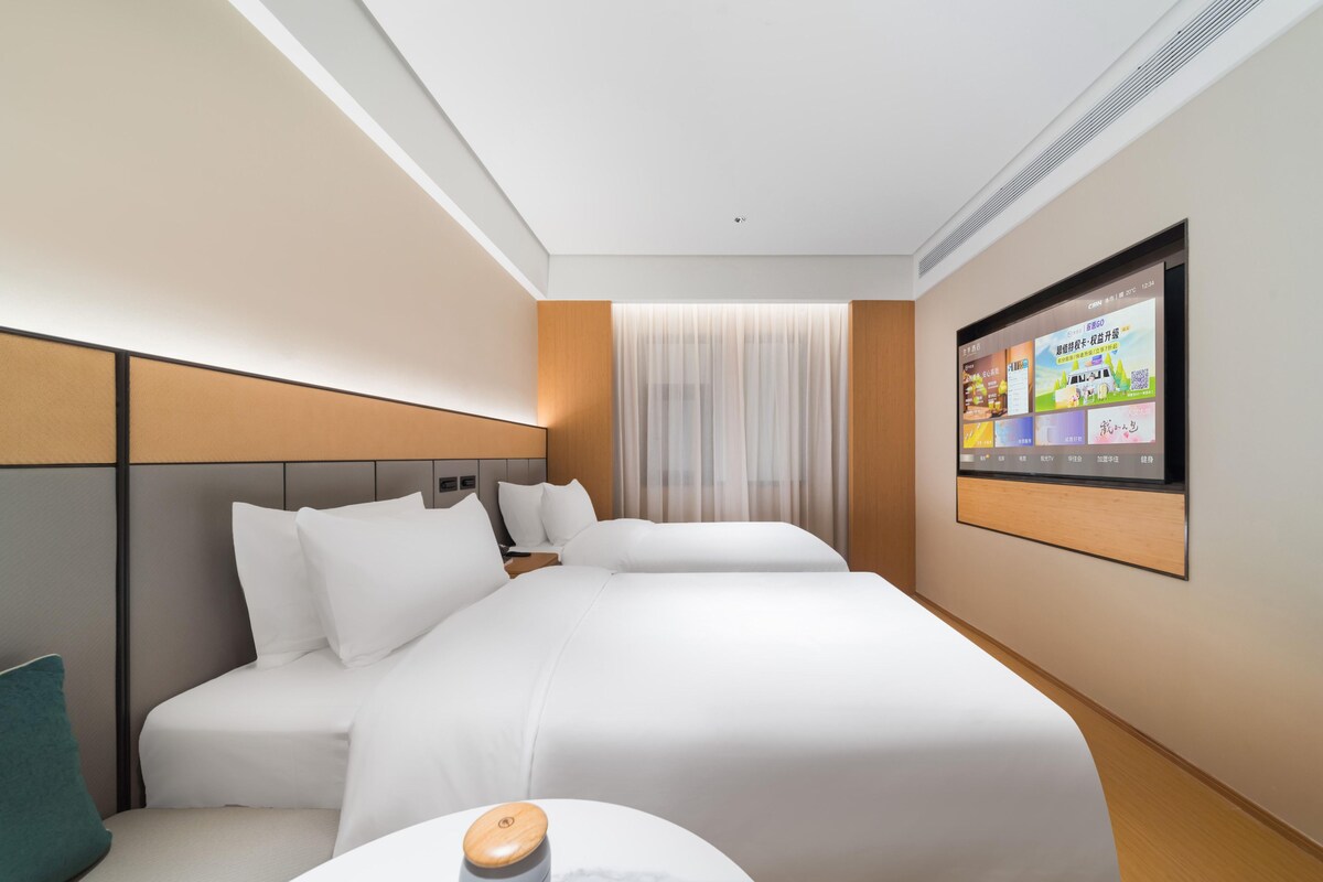 A hotel room with two beds, a TV mounted on the wall, and a window with sheer curtains.
