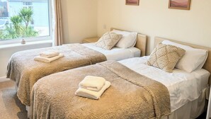 Ett hotellrum med två sängar, var och en med ett beige täcke och kuddar, ett fönster med utsikt över ett hus och en palm, och två inramade tavlor på väggen.