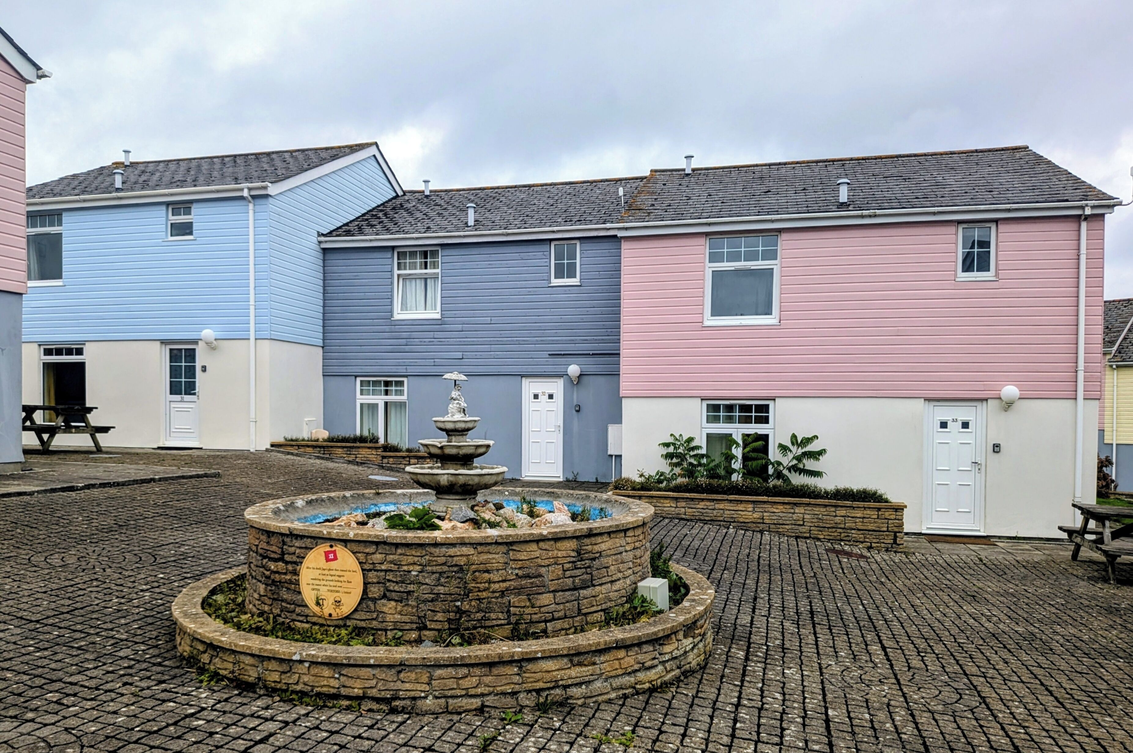 A courtyard with a stone fountain, a picnic table, and colorful houses.