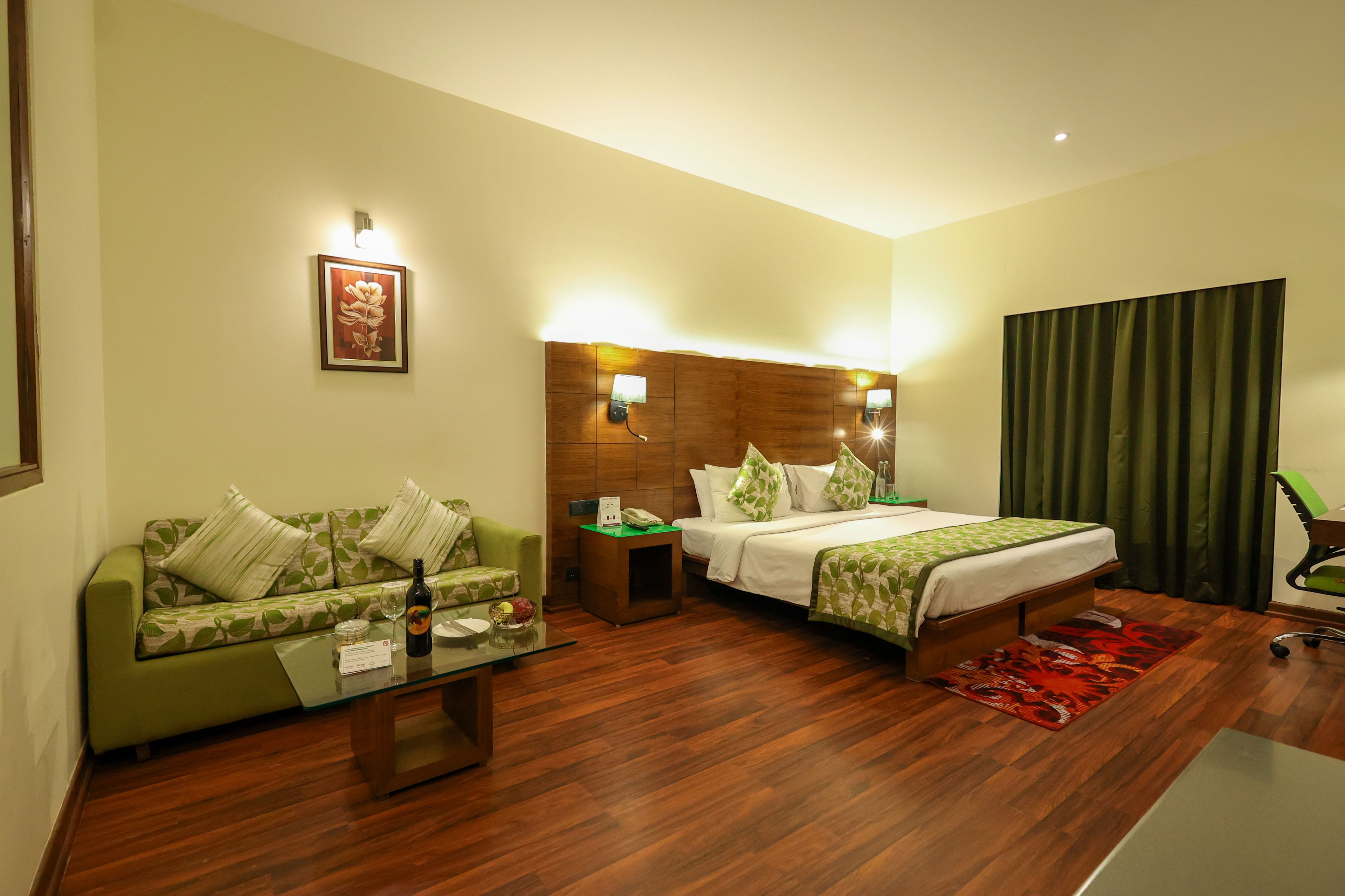 Habitación de hotel con un sofá verde, una cama con ropa de cama verde, un cabecero de madera, una mesa de centro de vidrio y una alfombra roja.