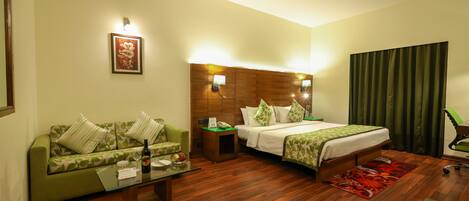 Habitación de hotel con un sofá verde, una cama con ropa de cama verde, un cabecero de madera, una mesa de centro de vidrio y una alfombra roja.