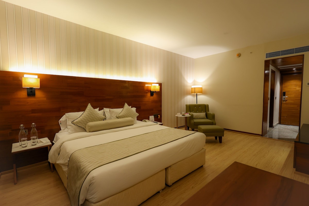 A hotel room with a large bed, bedside lamps, a small table, a green armchair, and a wooden headboard.