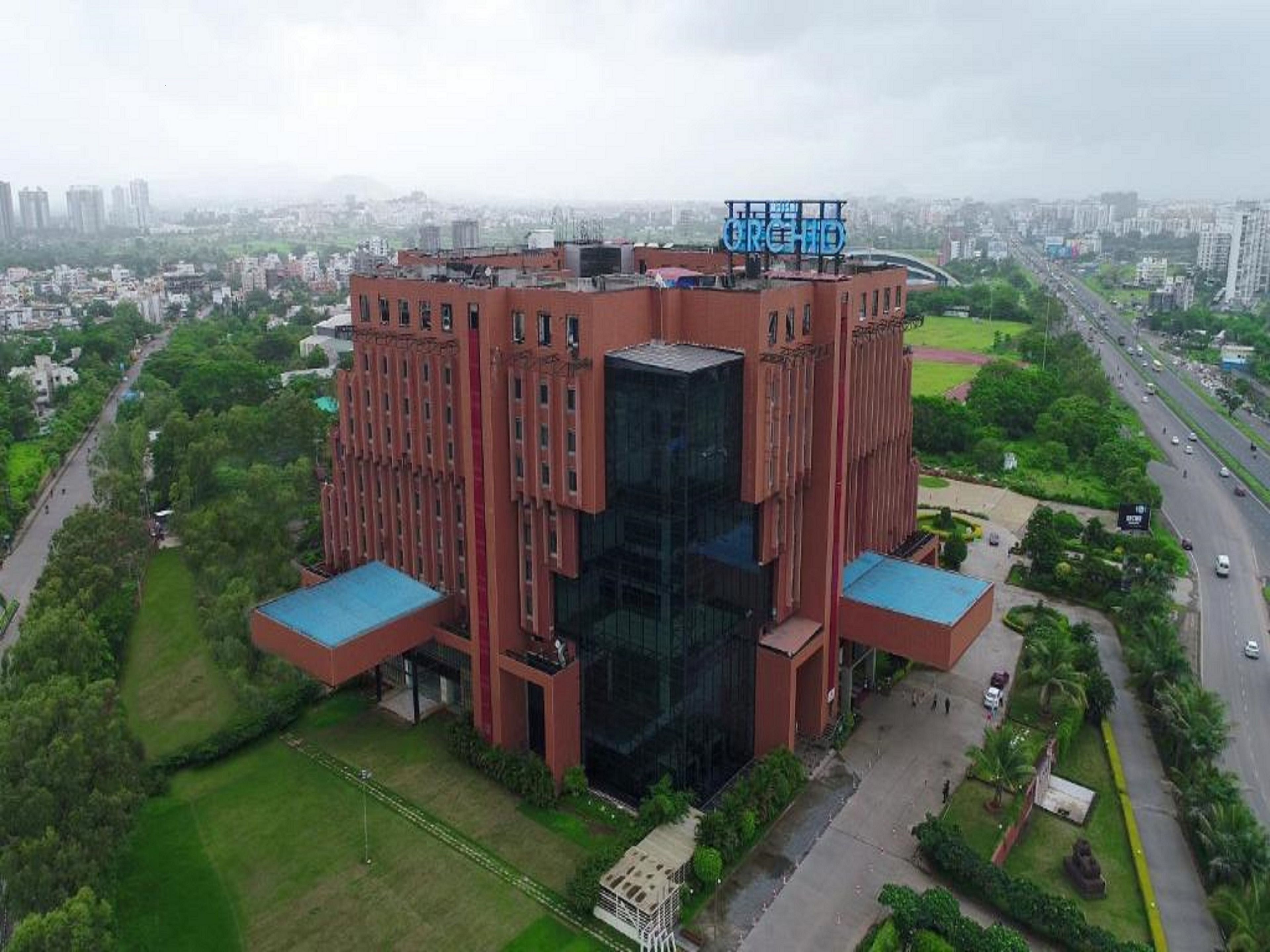A modern building with a glass facade and red brick exterior, surrounded by greenery and a busy road.