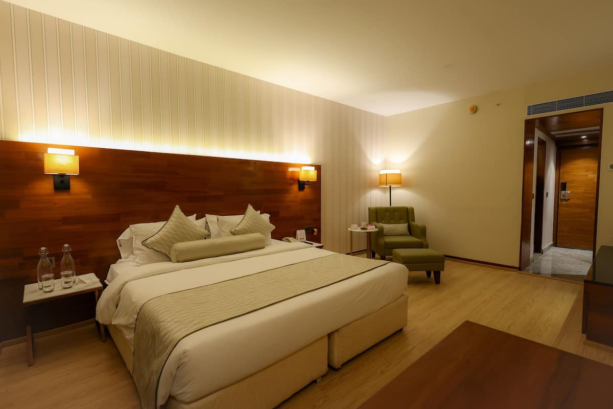 A hotel room with a large bed, two bedside lamps, a nightstand, a green armchair, and a wooden headboard.
