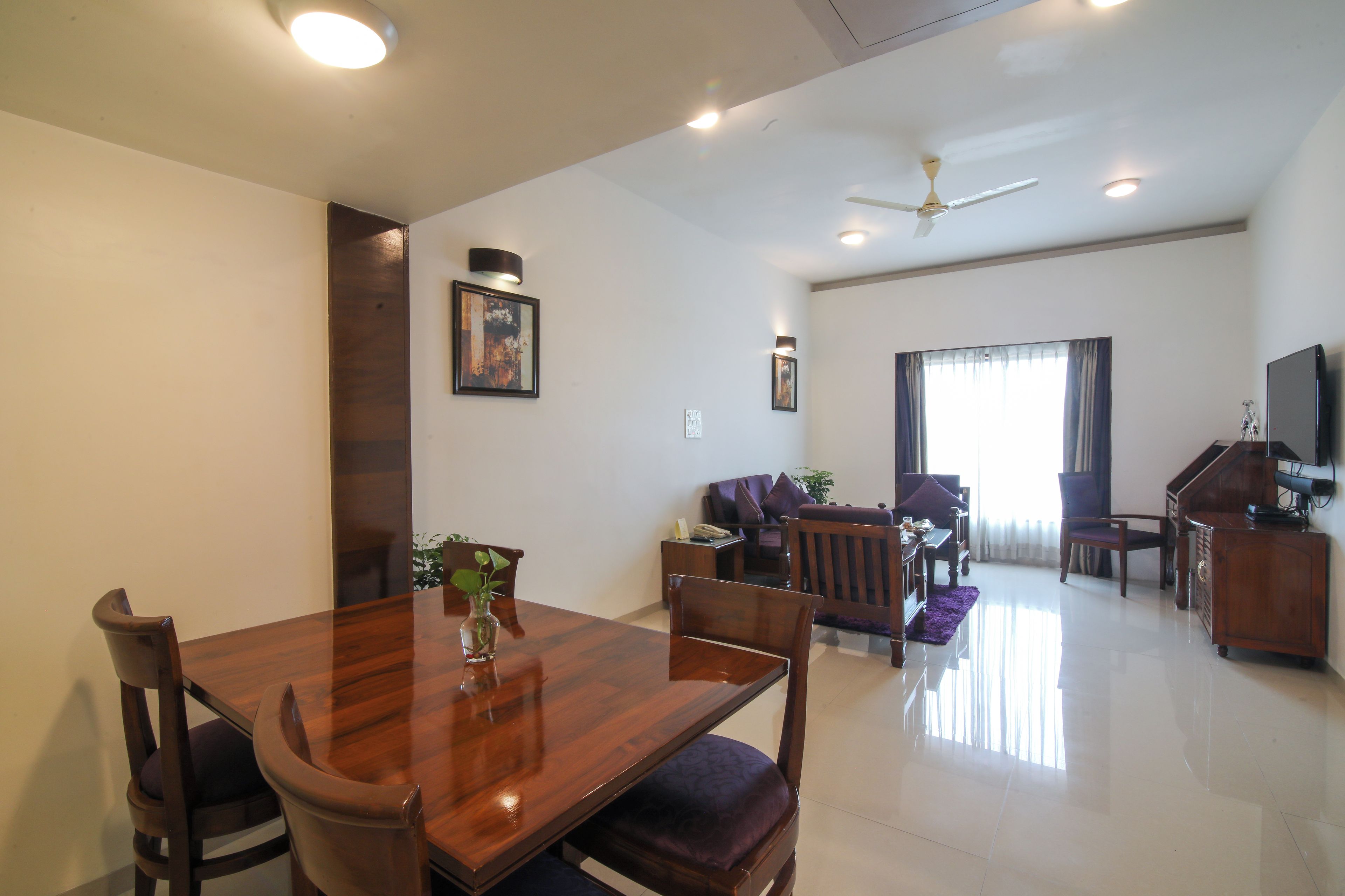 A spacious living room with a wooden dining table, comfortable seating, a ceiling fan, and a large window with curtains.