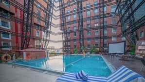 En swimmingpool med liggestol og håndklæde, omgivet af en bygning med rød murstensfacade og stillads.