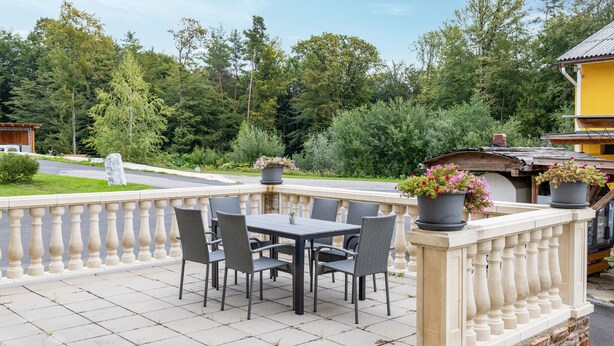An outdoor patio with a table and chairs, surrounded by a balustrade, with potted plants and a view of trees and a road. - Ferienwohnun Stainz 4 Personen (Stainz)