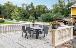Une terrasse extérieure avec une table et des chaises, entourée d’une balustrade, agrémentée de plantes en pot et offrant une vue sur des arbres et une route.
