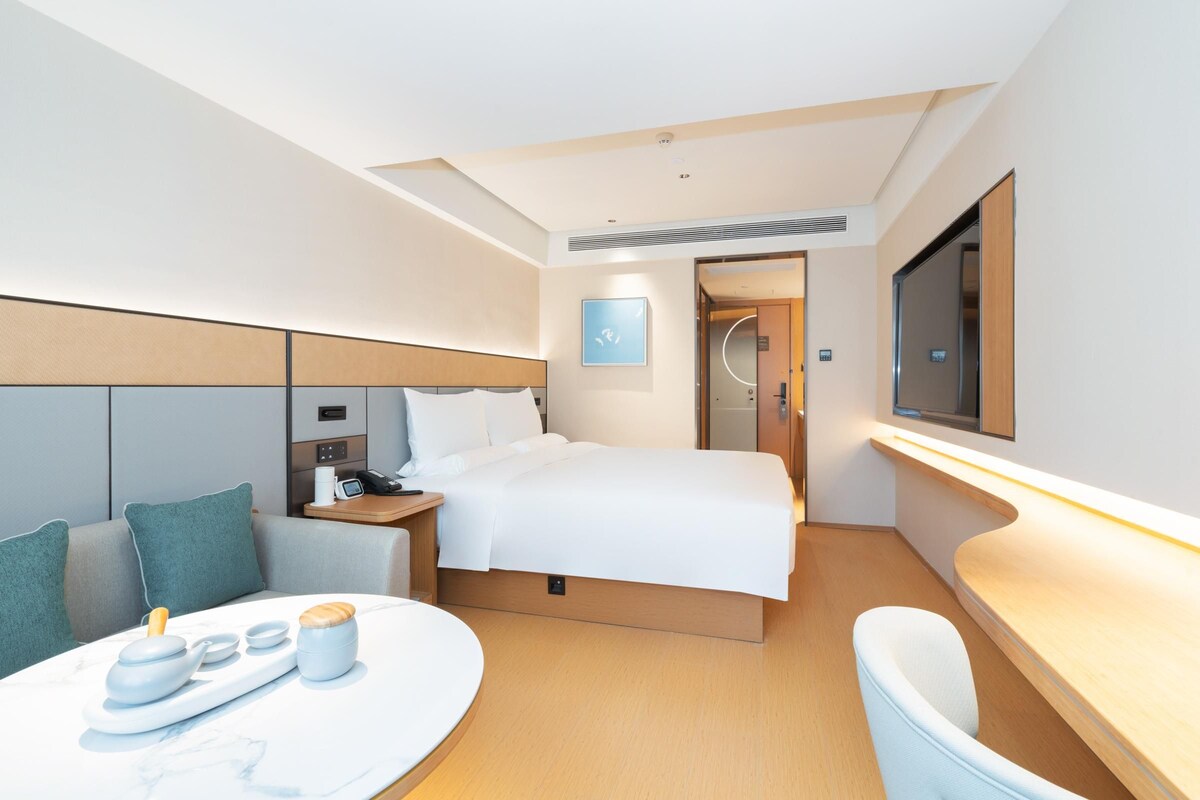 A modern hotel room with a bed, a desk, a seating area, and a television.