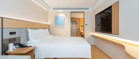 A modern hotel room with a neatly made bed, a bedside table with a phone and a coffee maker, a flat-screen TV mounted on the wall, and a large window providing natural light.