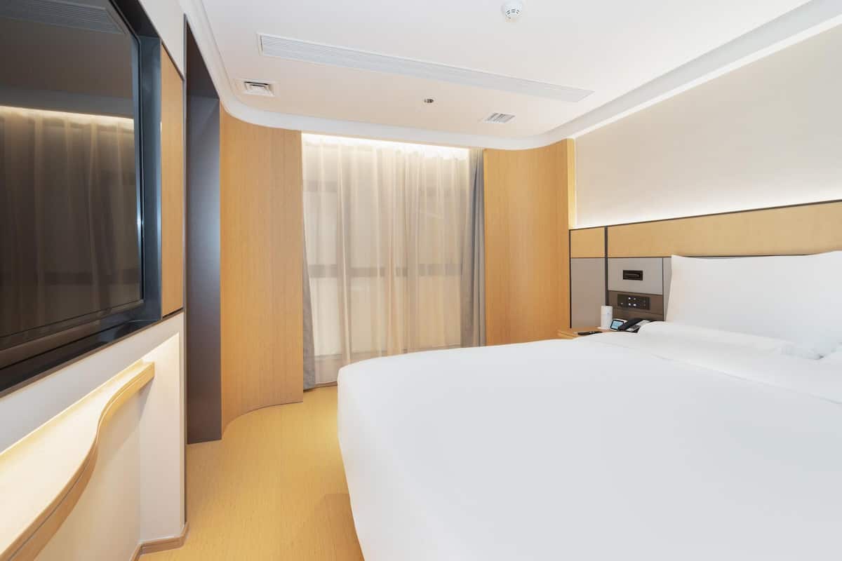 Una habitación de hotel moderna con una cama grande, una mesita de noche, televisor integrado y ventana con cortinas translúcidas.