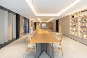 Une salle de conférence moderne, dotée d’une longue table en bois, de chaises blanches et d’une bibliothèque contenant des livres et des objets décoratifs.