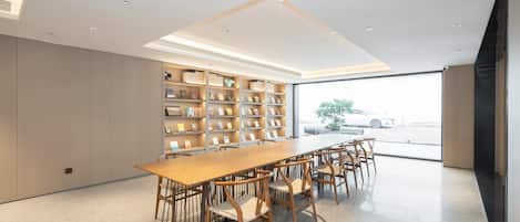 A modern library with a long wooden table and chairs, bookshelves, and a large window with a view of a car and trees.