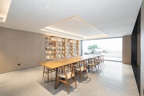 Une bibliothèque moderne avec une longue table et des chaises en bois, des étagères et une grande fenêtre donnant sur une voiture et des arbres.