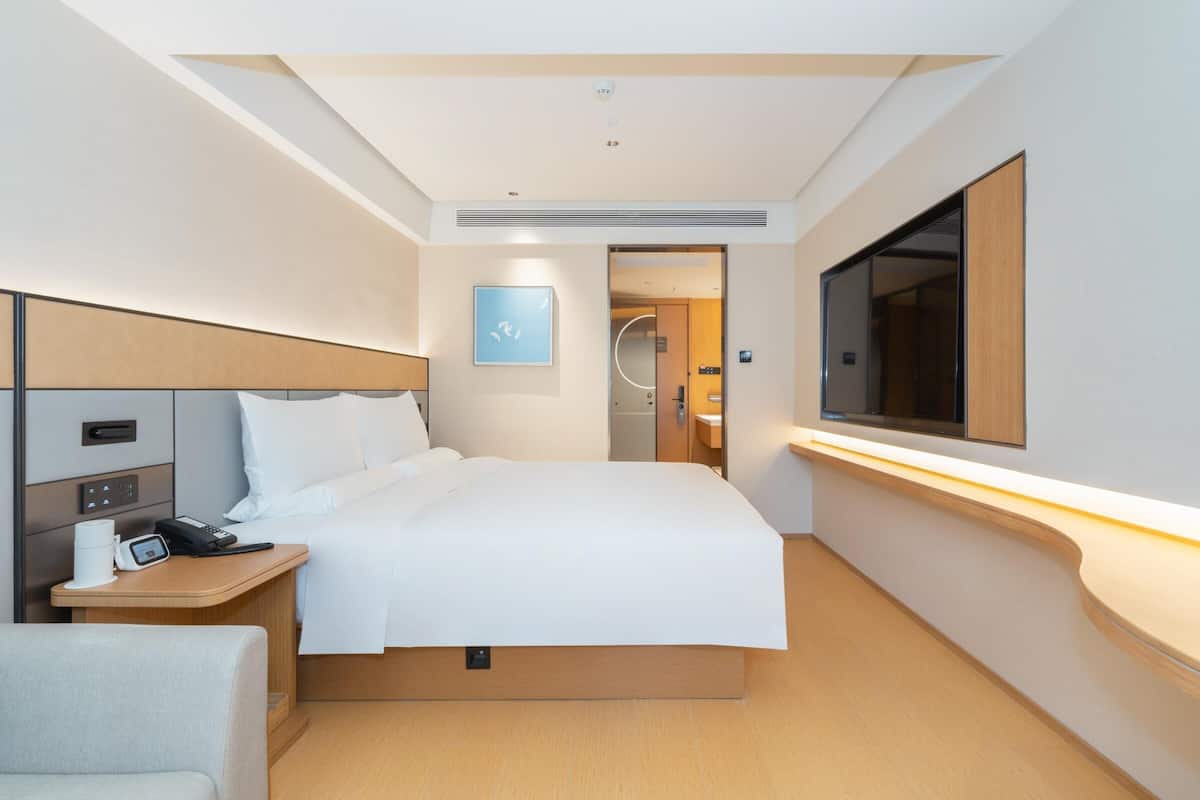 Una habitación de hotel moderna con una cama bien hecha, una mesita de noche con teléfono y cafetera, un televisor de pantalla plana montado en la pared y un amplio ventanal que proporciona luz natural.