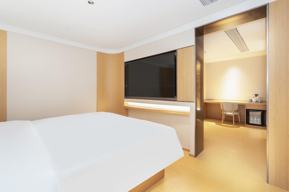 A modern hotel room with a large bed, a desk with a computer, and a small kitchenette.