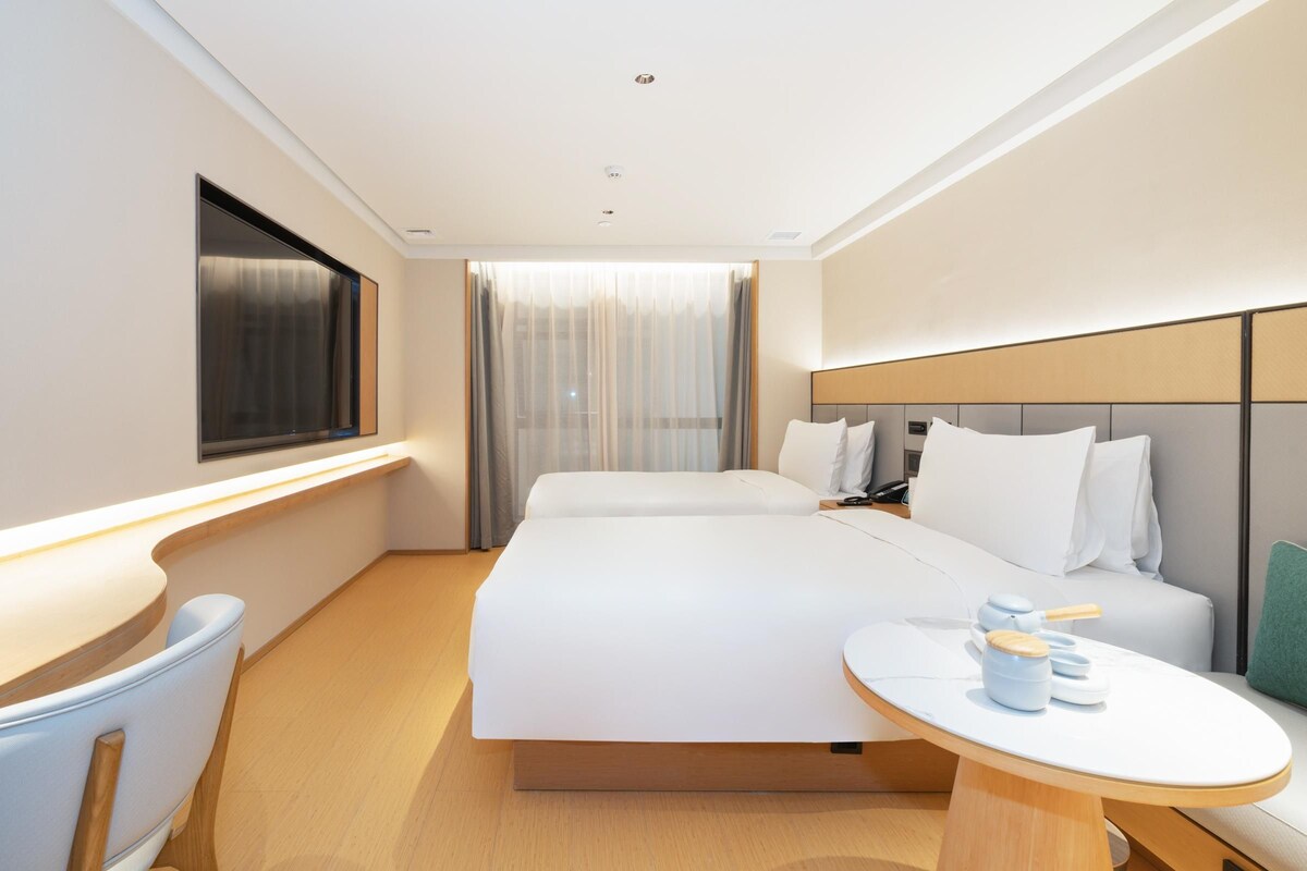 A modern hotel room with a large bed, bedside tables, a desk, and a window with curtains.