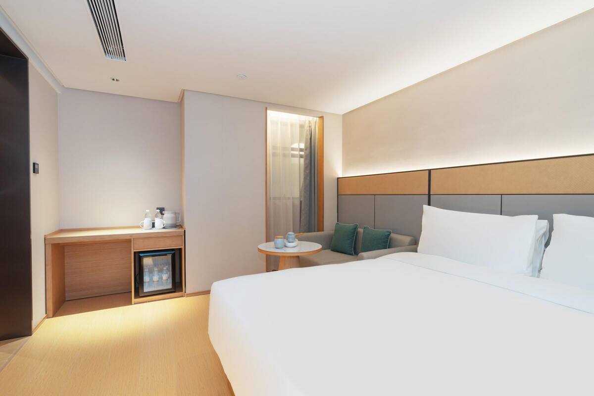 Una habitación de hotel moderna con una cama grande, un escritorio pequeño con refrigerador, una mesa redonda con una silla y un espejo amplio.