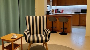 A living room with a striped armchair, a small wooden table with a cup, and a kitchen area with wooden cabinets and a bar stool. - Aegean Helios Houses (Rhodes)