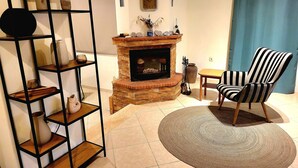 A living room with a brick fireplace, a striped armchair, and a black metal shelving unit with various decorative items. - Aegean Helios Houses (Rhodes)