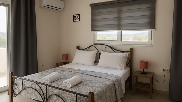 A bedroom with a bed, two bedside tables, a window with blinds, an air conditioning unit, and a view of the outdoors. - Aegean Helios Houses (Rhodes)