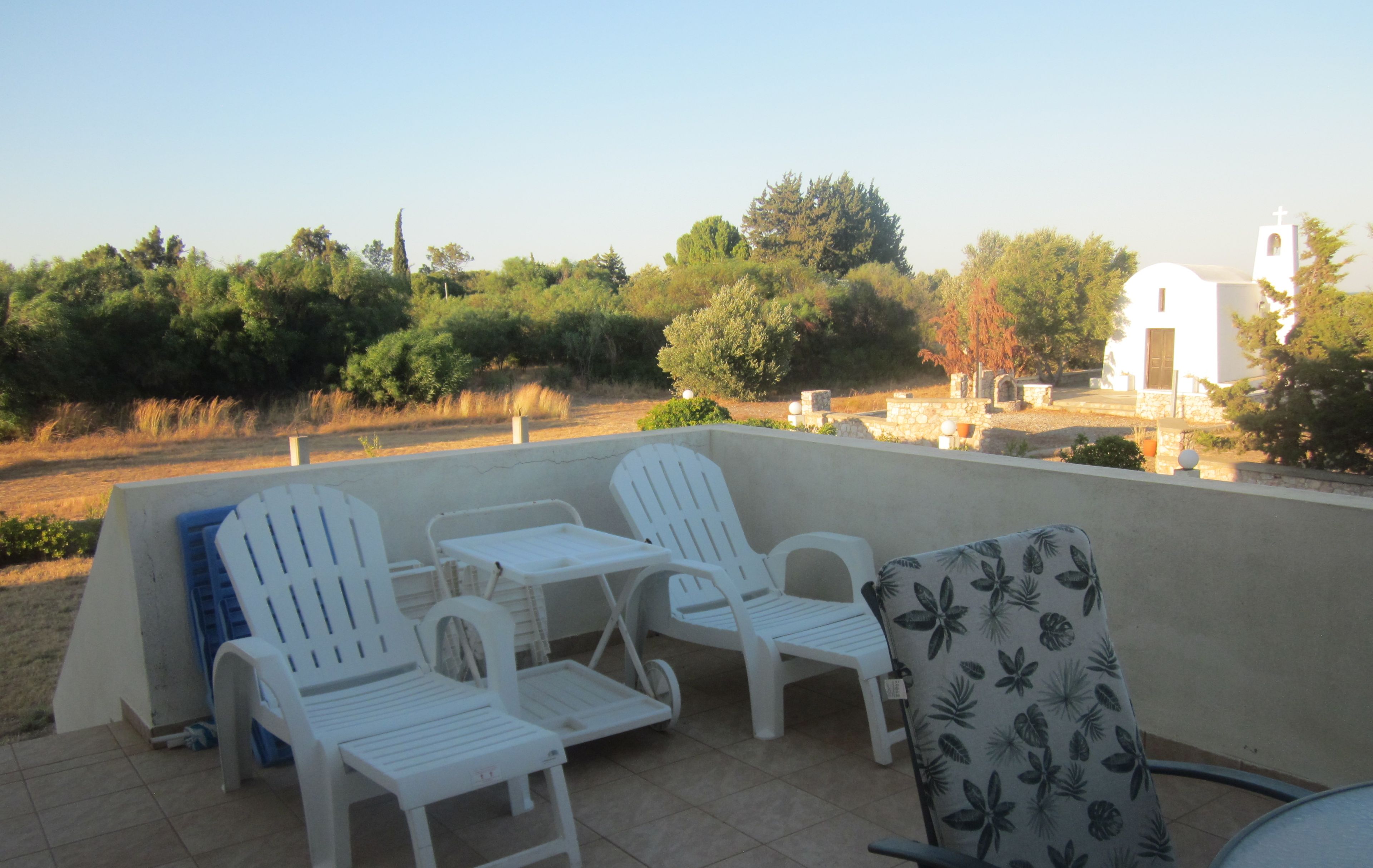 An outdoor patio with white plastic chairs and tables, a patterned chair, and a view of trees and a building in the background.