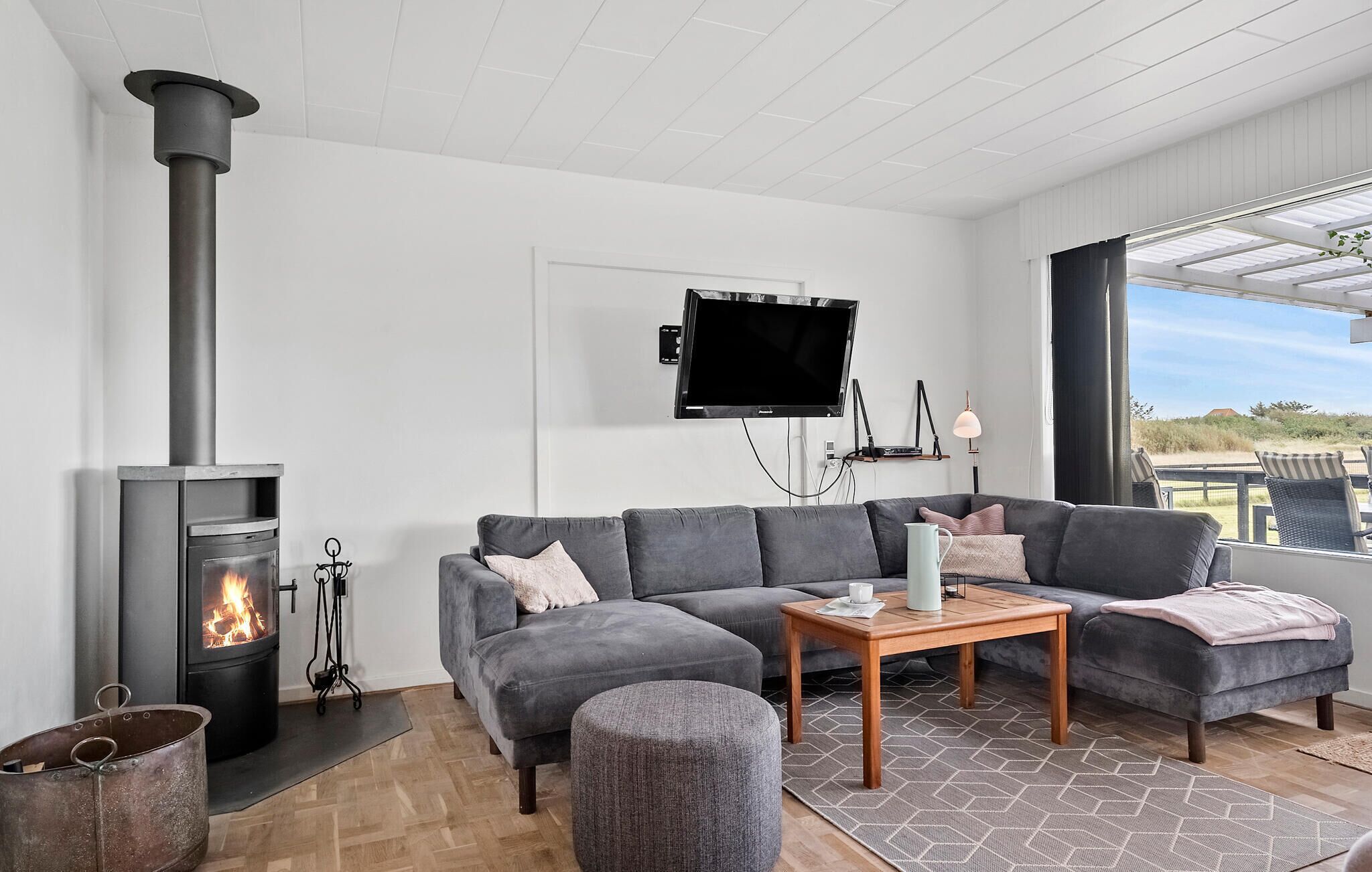 Un salon moderne doté d’un poêle à bois encastré, d’un canapé d’angle gris, d’une table basse en bois et d’une grande fenêtre offrant une vue sur l’extérieur.