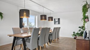 A dining area with a long wooden table, grey upholstered chairs, and black metal legs. There are three pendant lights with patterned shades hanging above. The room has white walls, a wooden floor, and large windows allowing natural light.