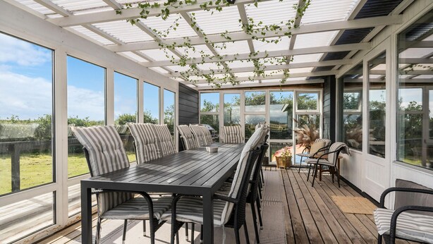 A covered patio with a wooden table and chairs, large windows, and a view of the outdoors.